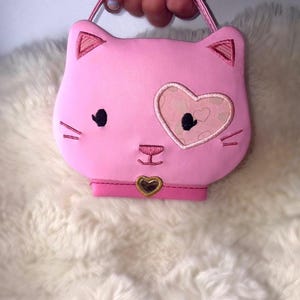 Puede incluir: Un bolso de mano rosa con forma de gato que presenta ojos negros bordados, un parche en forma de corazón sobre el ojo izquierdo y una nariz pequeña. El bolso incluye un collar rosa con un dije dorado en forma de corazón, dos orejas y un asa.