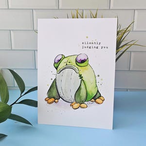 May include: A white greeting card featuring a watercolor illustration of a grumpy green frog with purple eyes. The text "silently judging you" is above the frog. The frog is sitting on a blue surface.
