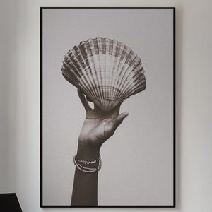 May include: A black and white photograph of a hand holding a large seashell. The hand is wearing a bracelet. The background is a light grey.