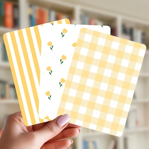 May include: Three decorative cards with rounded corners. The cards feature different patterns: yellow and white stripes, yellow flowers on white, and a yellow and white gingham check. The cards are held in front of a blurred bookshelf.