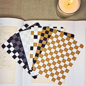 May include: A collection of checkered coasters in various colors, including black, white, and gold, are fanned out on an open book. A lit candle sits in the background, adding a warm ambiance to the scene. The coasters are square with rounded corners.
