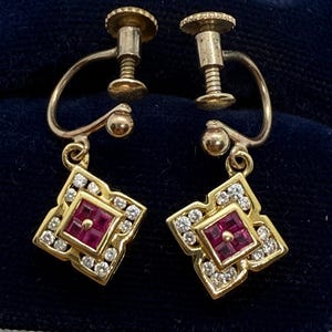 May include: A pair of gold screw-back earrings featuring a square-shaped design. Each earring contains a central square-cut red gemstone surrounded by a border of small, clear, round-cut white stones set within a gold frame with scalloped edges.