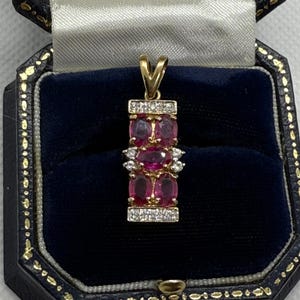 May include: A gold pendant with a rectangular design, featuring three oval-shaped, deep red gemstones. The pendant is accented with small, clear gemstones and is displayed in a blue velvet jewelry box.