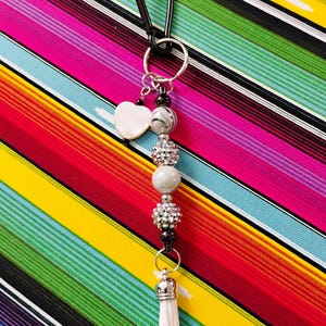 May include: A beaded keychain with a black carabiner clip. The keychain features a white heart charm, various beads in silver, gray, and black, and a small white tassel. The background is a colorful striped pattern.