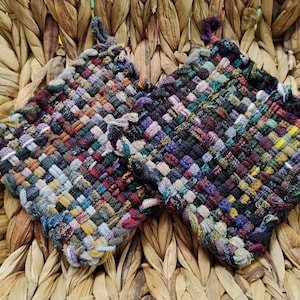 May include: Two square, woven potholders in a variety of colors including red, purple, green, and yellow. Each potholder has a loop for hanging. The potholders are sitting on a woven basket.