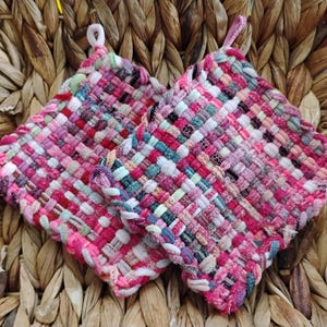 May include: Two square, woven potholders in shades of pink, red, and white, with small loops for hanging. The potholders are made of a woven fabric and are set against a woven background.