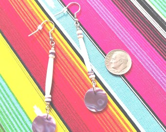 May include: A pair of dangle earrings featuring long white cylindrical beads, small dark spacers, and round purple marbled discs at the base. A United States dime is positioned next to the earrings to provide a clear sense of scale for the jewelry.