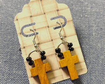 May include: A pair of dangle earrings featuring a wooden cross pendant in a natural tan finish, suspended from a silver-toned metal hook. Each earring includes a small circular wire accent adorned with four small black beads above the cross.