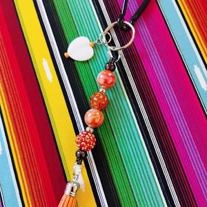 May include: A beaded keychain with a black carabiner clip. The keychain features a white heart charm, orange and red beads, and a small orange tassel. The background is a colorful striped pattern.