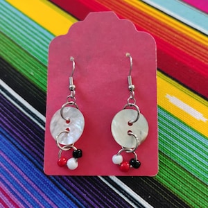 May include: A pair of dangle earrings featuring circular mother-of-pearl buttons attached to silver-toned metal hooks. Each earring includes a small silver ring dangling from the button, adorned with three tiny beads in solid red, white, and black.