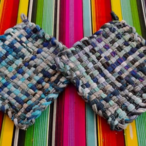 May include: Two square, woven potholders in shades of blue, gray, and white. Each potholder has a loop for hanging. The potholders are displayed on a colorful striped background.