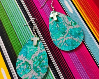 May include: A pair of teardrop-shaped earrings featuring a vibrant turquoise and green marbled pattern with a metallic silver overlay. Each earring is accented with a small, white, cross-shaped charm attached to a silver-toned metal hook wire.