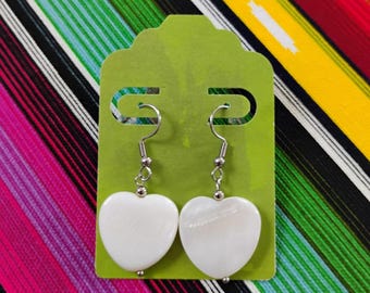 May include: A pair of white heart-shaped shell earrings with silver-toned metal hooks. The earrings are attached to a lime green rectangular display card with rounded top edges, featuring two horizontal slots for hanging the jewelry pieces.