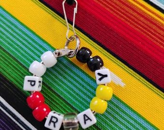 May include: A circular keychain featuring a silver metal clasp and ring strung with colorful plastic beads. The beads include white, black, red, and yellow spheres, along with white cube beads displaying the black letters P, R, A, Y.