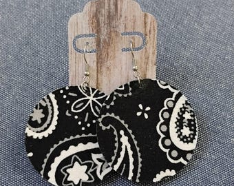 May include: A pair of circular drop earrings featuring a black, white, and gray paisley bandana print pattern. The earrings are attached to a light brown, wood-grain textured display card with two silver-toned metal earring hooks.