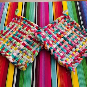 May include: Two colorful, woven potholders with loops for hanging. The potholders are made of various colors, including red, pink, yellow, green, and blue. They are placed on a striped, multi-colored surface.