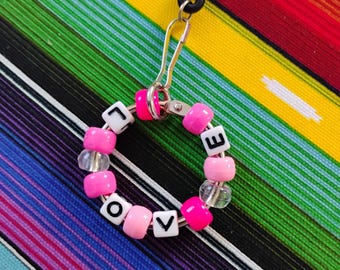 May include: A circular keychain featuring a silver metal ring adorned with alternating bright pink, light pink, and clear plastic beads. White square beads with the black letters L, O, V, and E are interspersed among the colorful beads. A black cord is attached to the ring via a silver metal hook.
