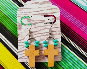 May include: A pair of dangle earrings featuring light brown wooden cross charms suspended from silver-toned metal hooks. Each earring includes a small silver ring accented with two bright turquoise beads on either side of the cross charm.