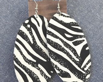 May include: A pair of oval-shaped drop earrings featuring a black and white zebra stripe pattern. The earrings are attached to a brown cardboard display card with silver-toned metal fishhook wires for pierced ears, showcasing a bold animal print.