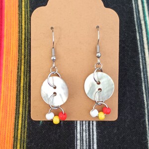 May include: Silver-tone button earrings with a dangling cluster of small beads in red, white, and yellow. The earrings are displayed on a brown card against a colorful striped textile background.