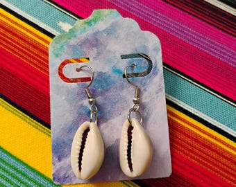 May include: A pair of dangle earrings featuring natural white cowrie shells suspended from silver-toned metal fishhook wires. The earrings are attached to a decorative display card with a blue and purple watercolor-style pattern and cutouts.