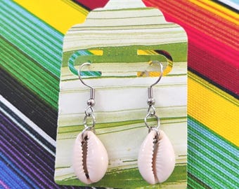 May include: A pair of dangle earrings featuring off-white cowrie shells suspended from silver-toned metal fishhook wires. The earrings are attached to a rectangular display card with a green and white marbled pattern and two die-cut slots.