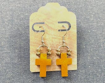 May include: A pair of drop earrings featuring mustard yellow wooden cross pendants suspended from silver-toned metal hooks. Each earring includes a small circular metal ring adorned with three tiny metallic beads above the cross charm.