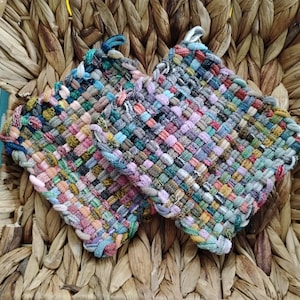 May include: Two handmade, square pot holders with woven, multicolored fabric. The pot holders feature loops for hanging and are set against a woven background. The colors include pink, blue, green, and yellow.