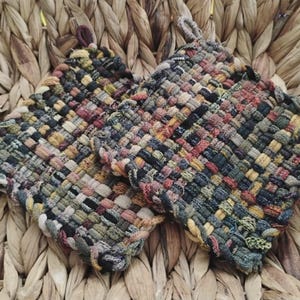 May include: Three woven potholders in shades of green, brown, and red, with a loop for hanging. The potholders are square-shaped and have a textured, woven appearance. They are resting in a woven basket.