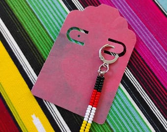 May include: A single dangle earring featuring a silver-toned hoop and a vertical charm made of two parallel strands of small seed beads. The beads are arranged in a color-blocked pattern of black, red, white, and yellow, attached to a pink display card.