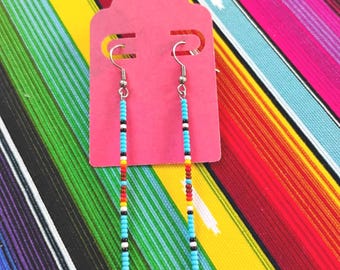 May include: A pair of long, slender dangle earrings featuring a vertical pattern of small seed beads in turquoise, white, black, red, and yellow. The earrings are attached to a bright pink rectangular display card with rounded top edges.