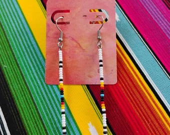 May include: A pair of long, slender dangle earrings featuring a vertical arrangement of small seed beads. The beads are primarily white, accented with a repeating pattern of black, red, yellow, and turquoise blue beads along the length of each earring.