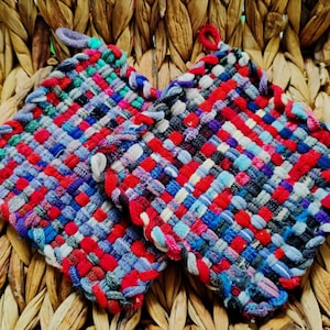 May include: Two square, woven potholders in a basket weave pattern. The potholders are primarily red, blue, and white, with accents of purple, green, and black. Each potholder has a small loop for hanging. The potholders are sitting on a woven wicker surface.