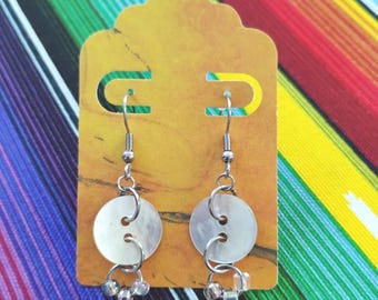 May include: A pair of dangle earrings featuring circular white mother-of-pearl buttons suspended from silver-toned metal hooks. Each earring includes a small silver jump ring connecting the button to three tiny, clear, translucent glass beads.