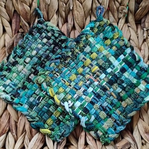 May include: Two square, woven potholders in shades of green, blue, and yellow. Each potholder has a loop for hanging. The potholders are made of a tightly woven material, and are displayed on a woven background.