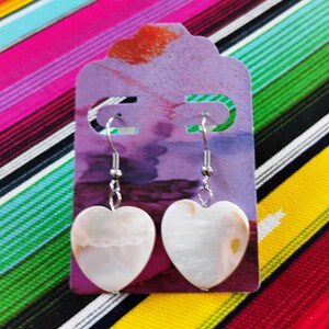 May include: Heart-shaped earrings with a pearlescent finish hang from silver-colored hooks. The earrings are displayed on a purple card with a decorative top, set against a colorful striped background. The earrings are a jewelry item.