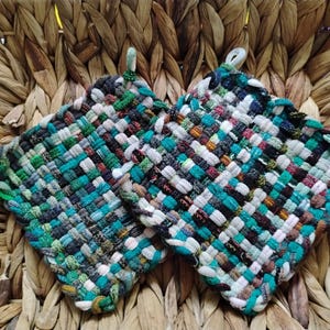 May include: Two square, woven potholders in shades of teal, green, gray, and white. Each potholder has a loop for hanging. The potholders are made of a woven fabric and are set against a woven background.