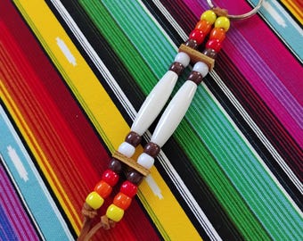 May include: A keychain featuring two parallel leather cords strung with yellow, orange, red, brown, and white beads, accented by two long white cylindrical beads in the center, finished with a silver metal ring at the top and loose leather ends.