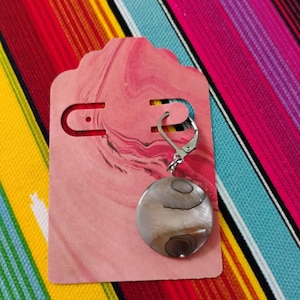 May include: A single circular earring featuring a polished, iridescent shell disc with natural brown and grey swirling patterns, attached to a silver-toned metal lever-back finding, displayed on a pink marbled rectangular card with rounded edges.