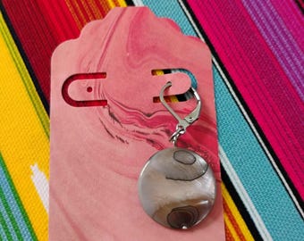 May include: A single circular earring featuring a polished, iridescent shell disc with natural brown and grey swirling patterns, attached to a silver-toned metal lever-back finding, displayed on a pink marbled rectangular card with rounded edges.