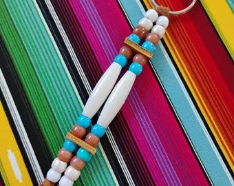 May include: A keychain featuring two parallel strands of beads attached to a metal ring. The design includes white, brown, and turquoise round beads, two long white cylindrical beads, and two small tan leather spacers, ending in two leather cord tails.