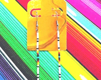 May include: A pair of long, slender dangle earrings featuring a repeating pattern of small seed beads in white, black, red, yellow, and turquoise. The earrings are suspended from silver-toned metal hooks and displayed on a yellow card.