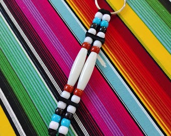 May include: A keychain featuring two parallel strands of beads attached to a metal ring. The design includes white, turquoise, black, and rust-colored beads, along with two long, smooth white cylindrical beads in the center, all secured by black cord.