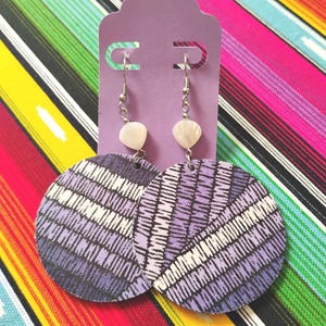 May include: A pair of circular drop earrings featuring a purple and white striped pattern. Each earring hangs from a silver-toned metal hook and includes a small, white, shell-like accent bead positioned above the circular disc component.