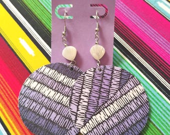 May include: A pair of circular drop earrings featuring a purple and white striped pattern. Each earring hangs from a silver-toned metal hook and includes a small, white, shell-like accent bead positioned above the circular disc component.