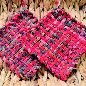 May include: Two handmade, square potholders with woven red, pink, and blue fabric. Each potholder has a small loop for hanging. The potholders are displayed on a woven wicker background.