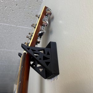 May include: A black guitar wall mount with a geometric design holds a wooden acoustic guitar with chrome tuning pegs.