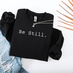 May include: Black sweatshirt with the text "Be Still." printed in white on the front.