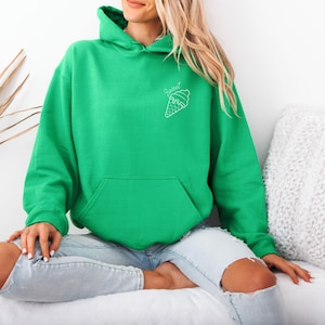 Op de afbeelding: Een felgroene hoodie met een voorzak en een capuchon. De hoodie heeft een witte ijsje-afbeelding met het woord "Sweet" erboven. Het model draagt een lichtblauwe jeans.