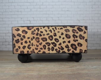 Puede incluir: Una caja de madera rectangular con un diseño de estampado de leopardo. La caja tiene patas de madera negras y está sentada sobre una superficie de madera.